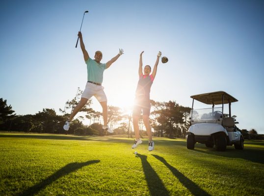 Full length of golf player couple with arms raised while jumping on field