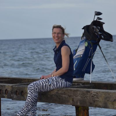Franca Fehlauer mit Golfschlägern am Meer sitzend und lachend